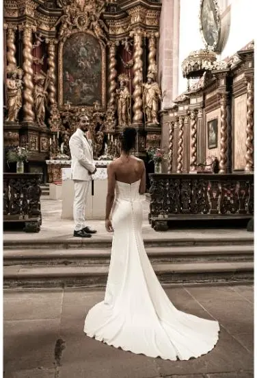 Ein Bräutigam in einem stilvollen weißen Anzug steht in einer prachtvoll dekorierten Kirche, während die Braut in einem eleganten weißen Hochzeitskleid den Mittelgang entlangschreitet.