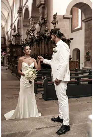 Das Brautpaar steht in einer historischen Kirche, der Bräutigam hält sanft die Hand der Braut, die ein strahlendes Lächeln zeigt und einen weißen Blumenstrauß trägt.
