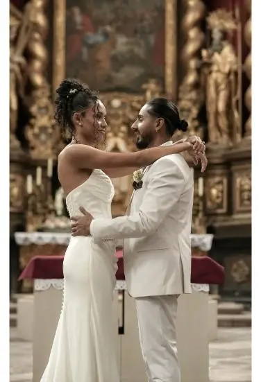 Das Brautpaar umarmt sich innig vor einem detailreich verzierten Altar, beide tragen stilvolle weiße Hochzeitskleidung.