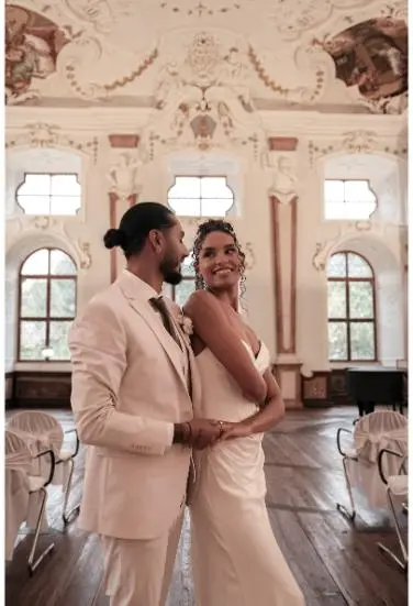 Das Brautpaar steht eng umschlungen in einem prächtigen, barock dekorierten Saal mit hohen Fenstern und einer kunstvollen Decke.