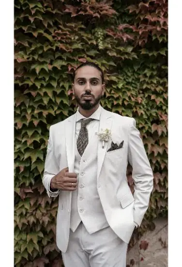 Ein Bräutigam in einem weißen Anzug steht selbstbewusst vor einer mit Efeu bewachsenen Wand und schaut direkt in die Kamera.