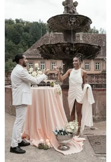 Das Brautpaar steht an einem dekorativen Brunnen und stößt mit Champagnergläsern an, die Braut trägt ein kurzes, modernes Hochzeitskleid.