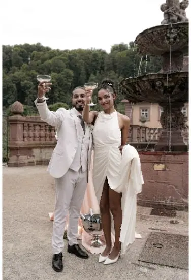 Die Braut und der Bräutigam heben ihre Champagnergläser und lächeln fröhlich in die Kamera, der Hintergrund zeigt einen historischen Außenbereich.