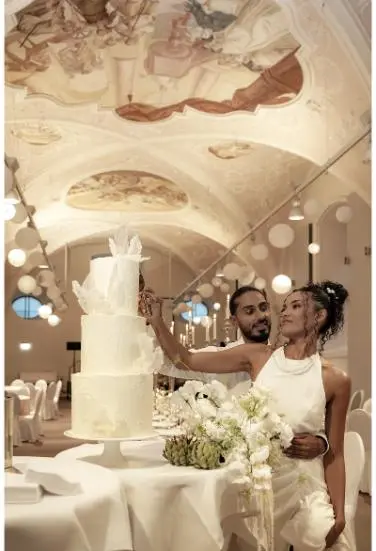 Das Brautpaar schneidet gemeinsam eine dreistöckige, elegante Hochzeitstorte in einem festlich dekorierten Saal mit verzierten Decken an.