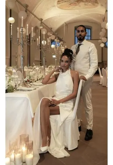 Das Brautpaar posiert an einer festlich gedeckten Hochzeitstafel; die Braut sitzt elegant, während der Bräutigam hinter ihr steht.