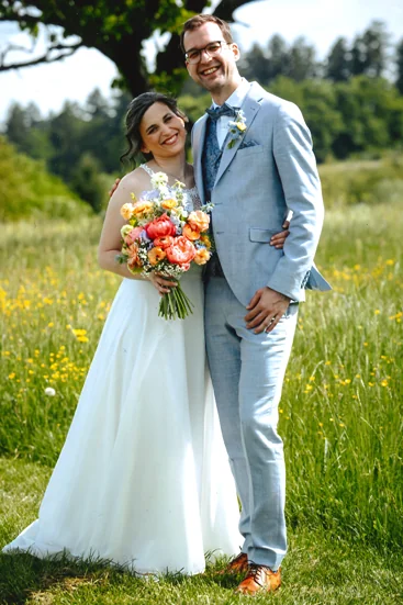 Glückliches Brautpaar in einer Sommerwiese – die Braut im weißen Kleid mit buntem Blumenstrauß, der Bräutigam im hellblauen Anzug mit Fliege, beide lächelnd und Arm in Arm.