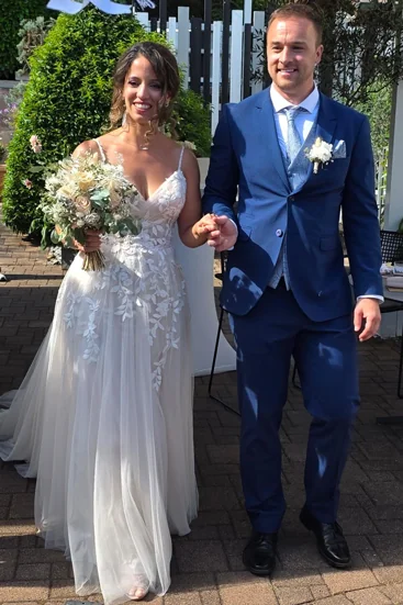 Frisch verheiratetes Brautpaar beim Auszug – die Braut in einem luftigen Spitzenkleid mit floralen Details, der Bräutigam in einem blauen Hochzeitsanzug mit Weste und Ansteckblume. Beide strahlen und halten sich an den Händen.