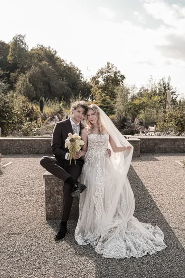 Braut und Bräutigam posieren auf Steinbank, beide schauen in die Kamera